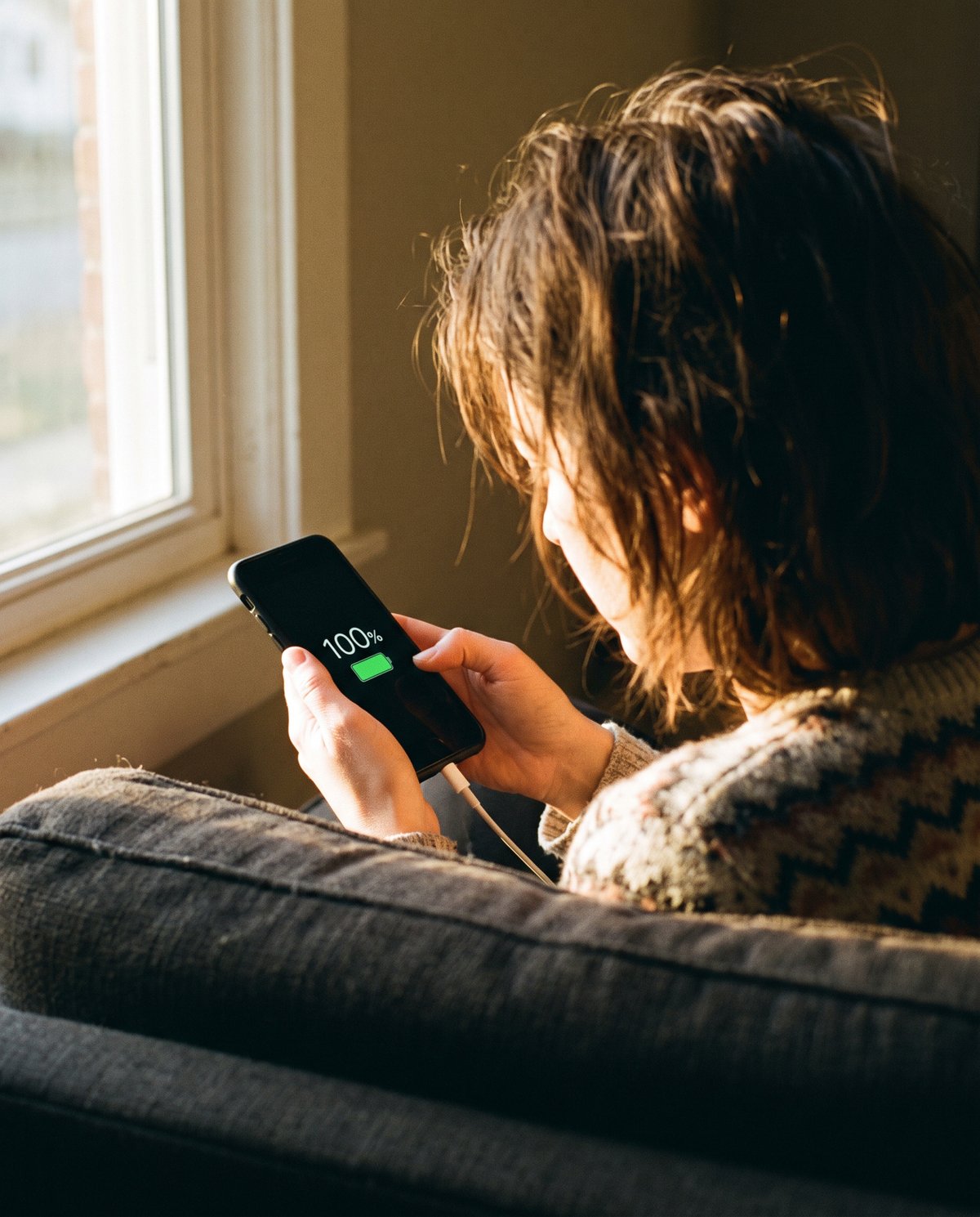Over her shoulder: a woman on a sofa looks at her charging phone, which shows 100% and a full battery.